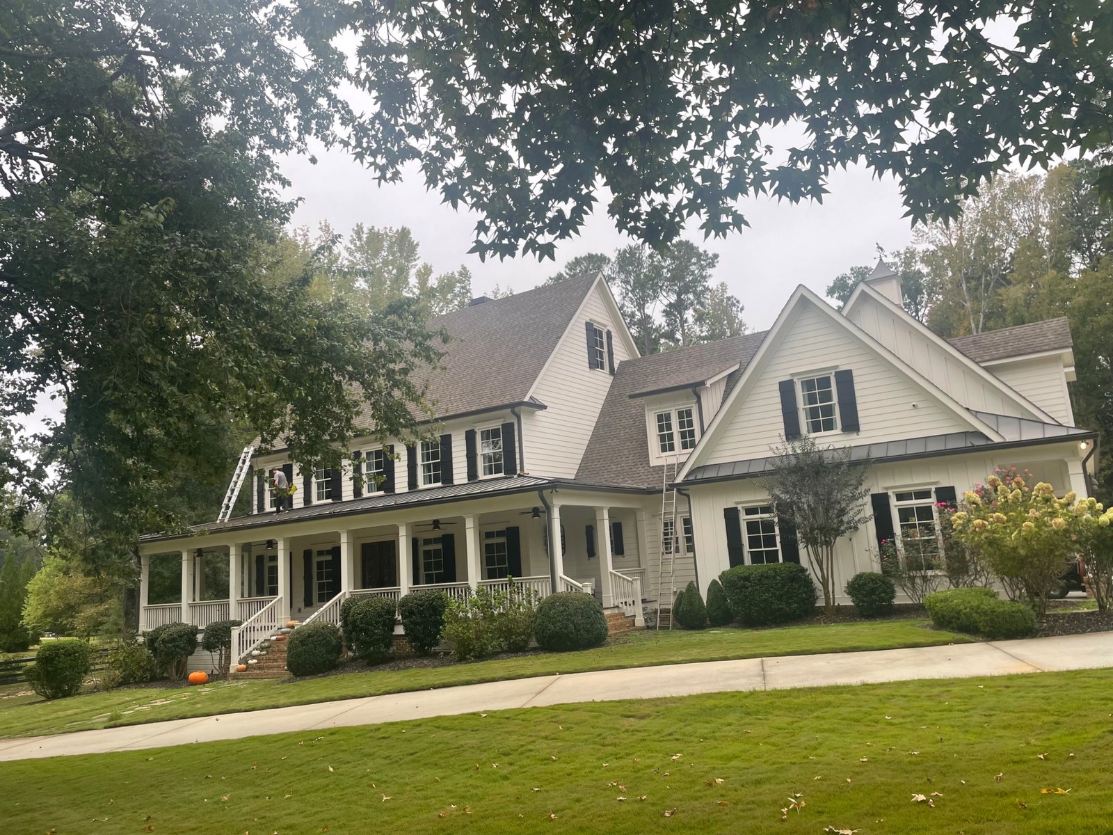A ladder set against a two-story white house with a covered front porch, for gutter cleaning and wood repairs